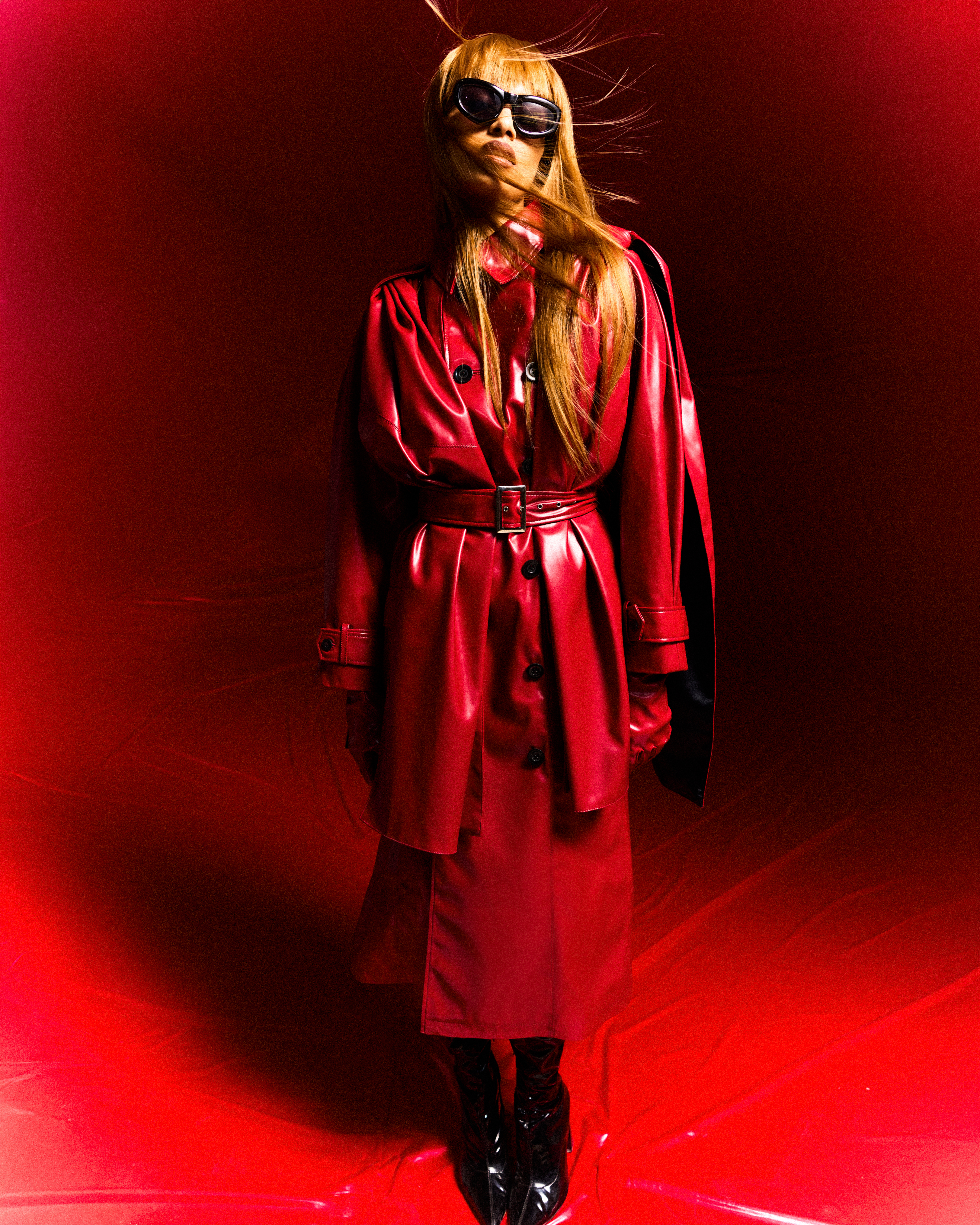 Fashionable woman in a red vinyl trench coat, wearing sunglasses and gloves, stands against a matching red backdrop in a studio setting.