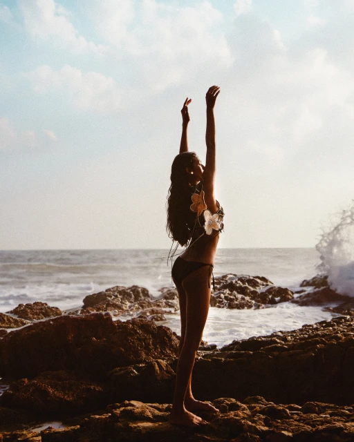Woman in bikini poses with arms raised on rocky beach with ocean waves and sky. The light is warm, creating a summery outdoor scene.