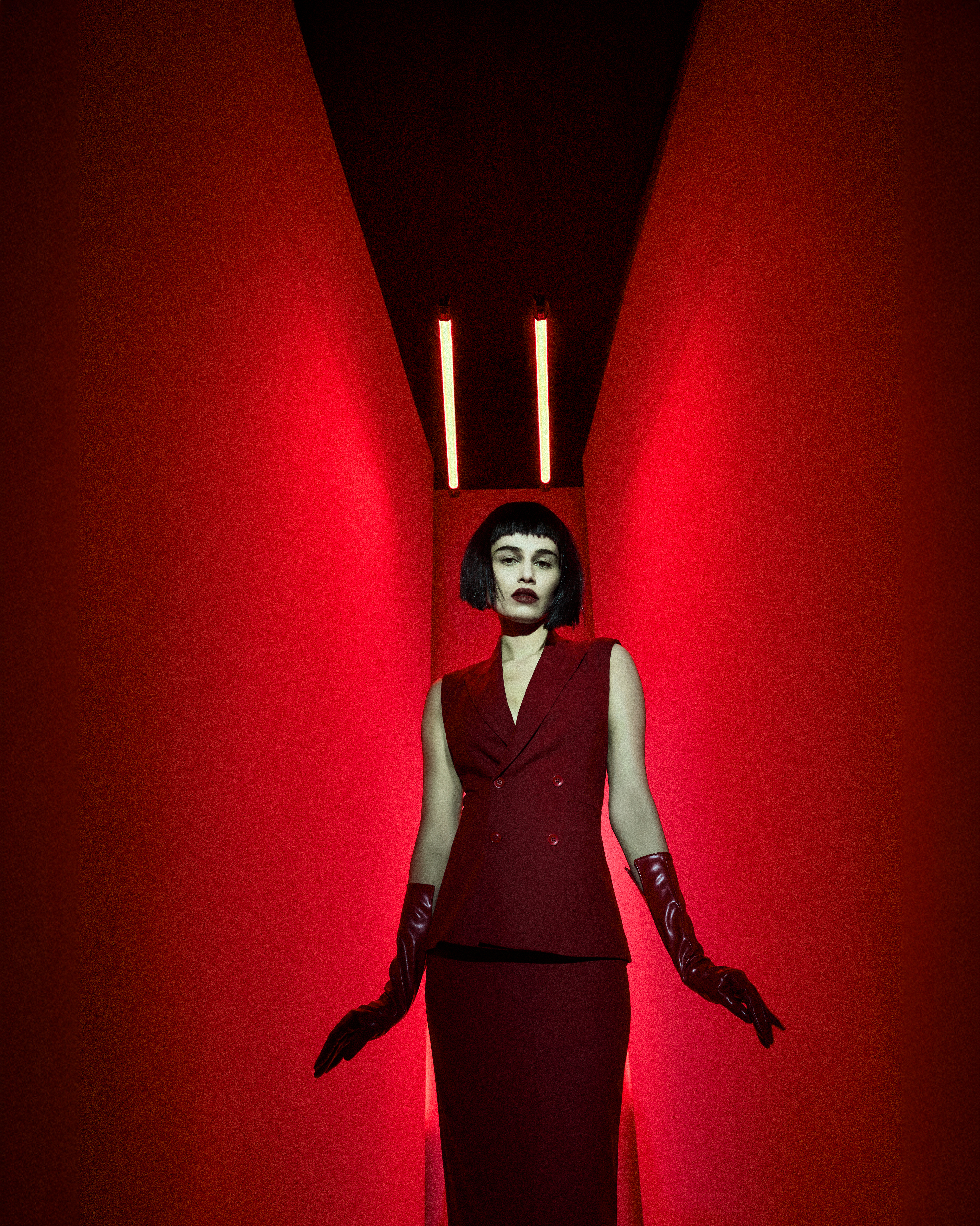 A woman in a red vest and skirt stands in a red hallway under two lights. She wears long red gloves and has short dark hair with bangs.
