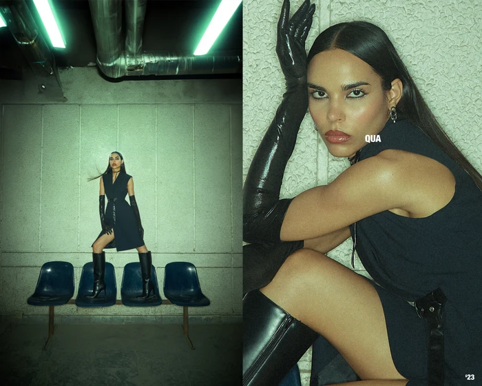 Woman in dark sleeveless dress wearing long black gloves and knee-high boots posing on chairs under a green-lit industrial ceiling.