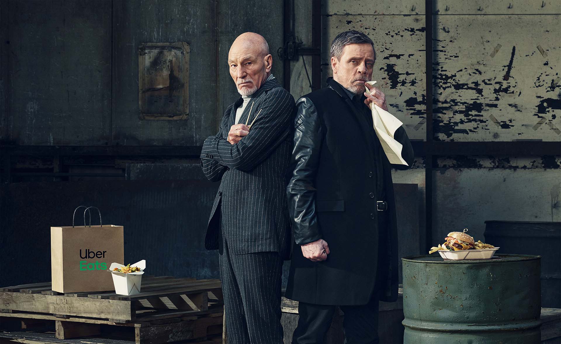 Two adult males in suits stand back-to-back in a gritty urban setting. Food delivery items are placed in the foreground.