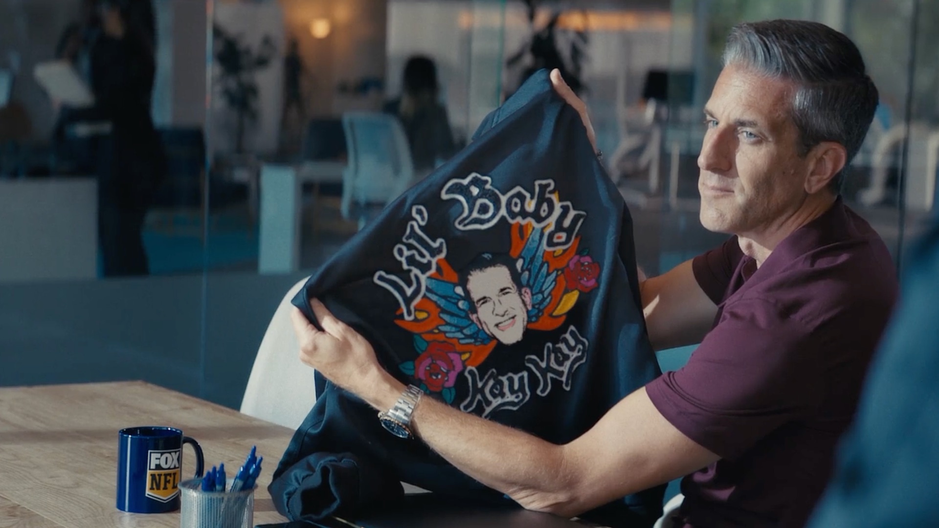 Adult male sitting at a conference table in an office space, holding up a black jacket with colorful graphics; Fox NFL mug on table.