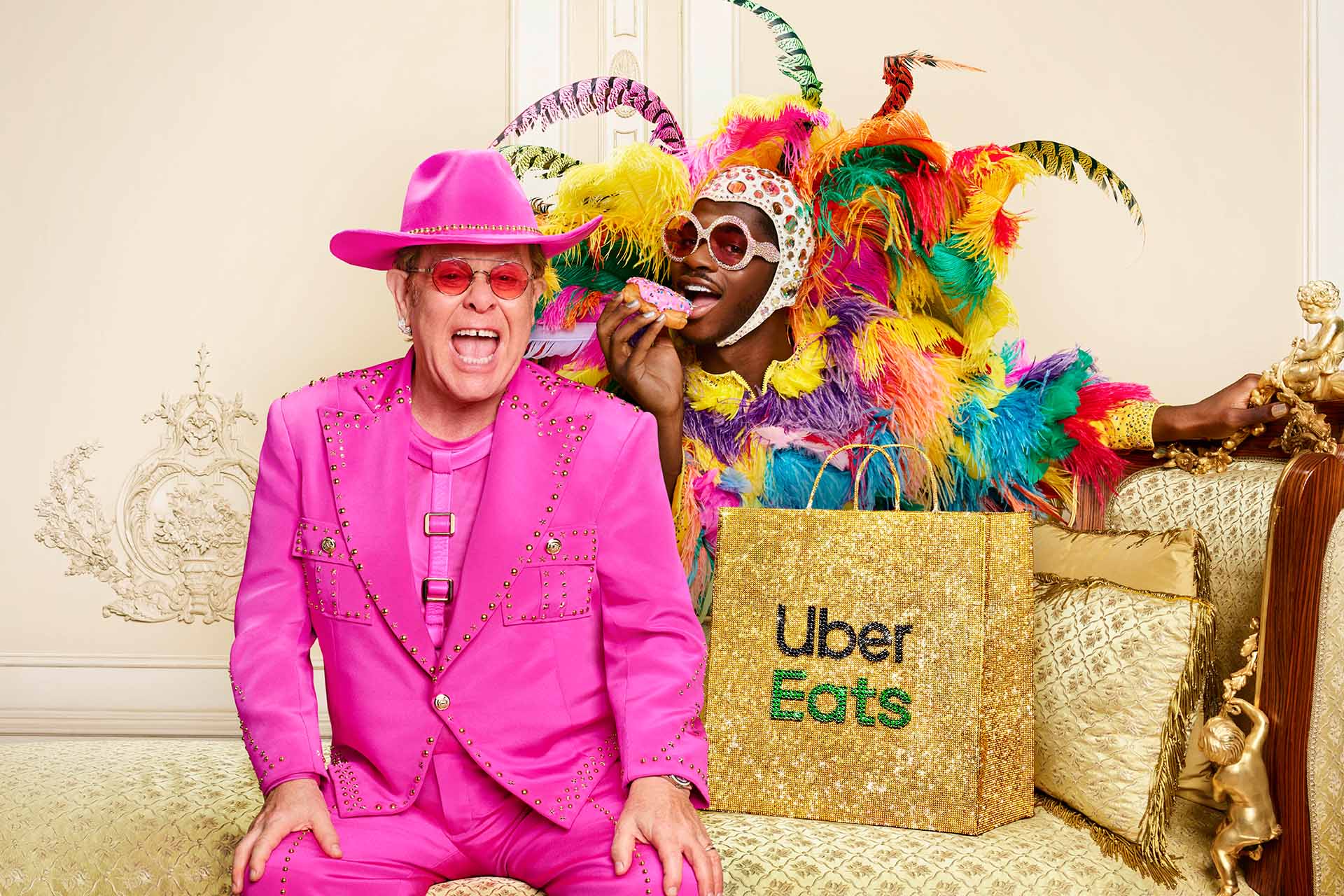Elton John in a pink suit with Lil Nas X eating a donut in colorful feathers for Uber Eats, sitting on a gold accented couch.