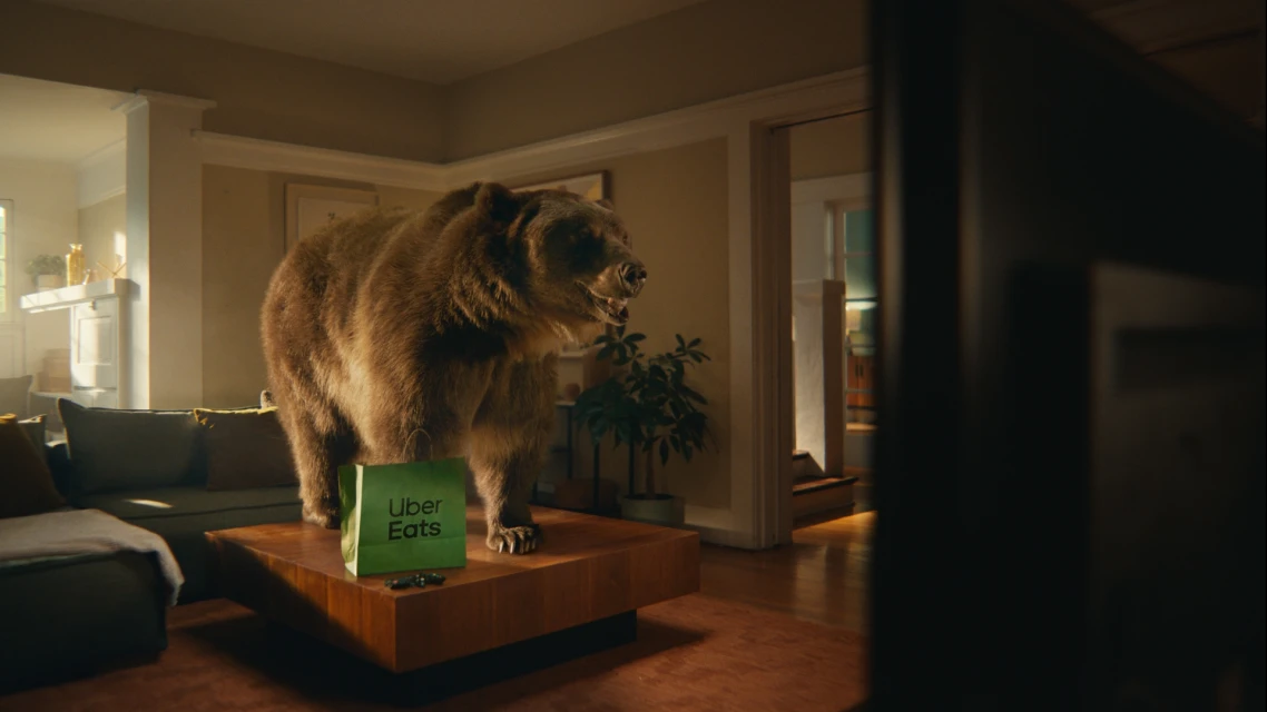 A brown bear stands on a coffee table with an Uber Eats bag, in a living room with a TV, creating a humorous and unexpected scene.