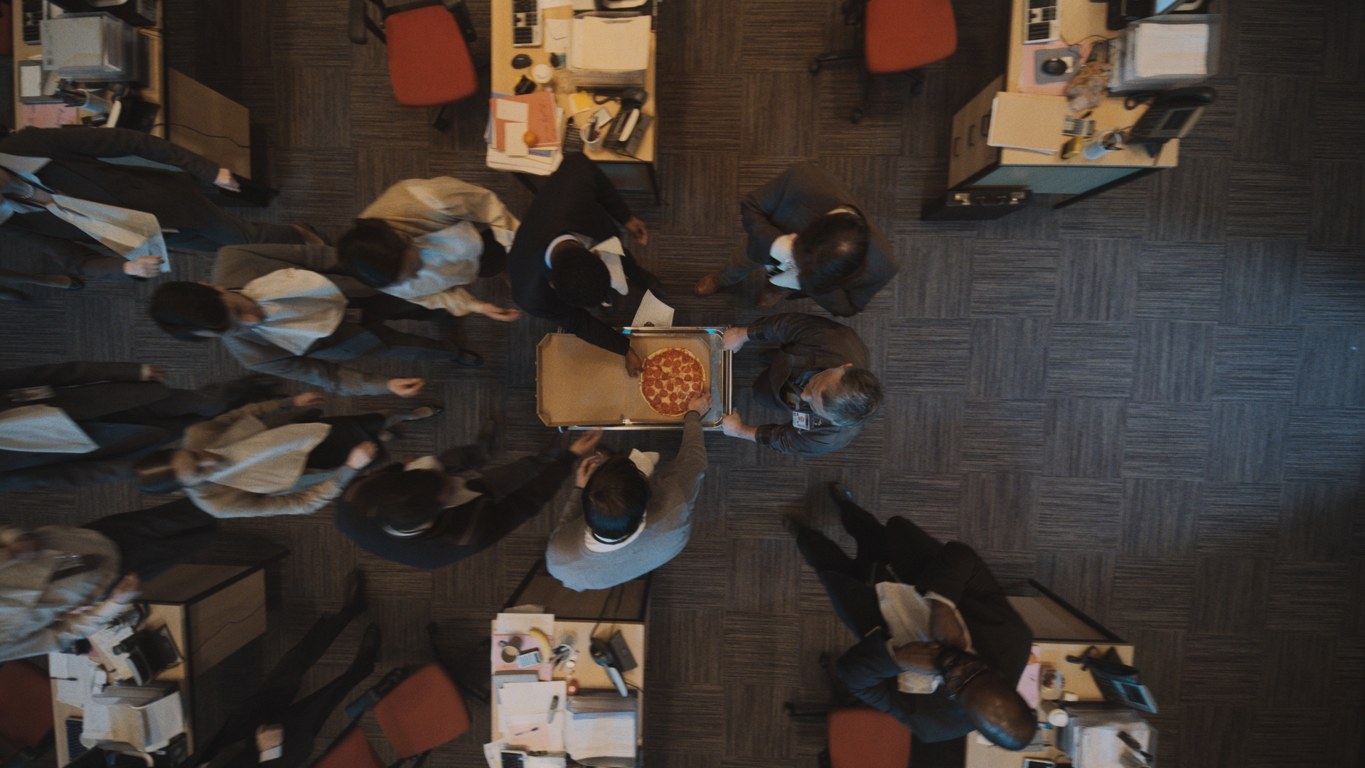 Overhead shot of diverse office workers gathering around a pizza in a box, set on a wheeled cart in a modern office setting.