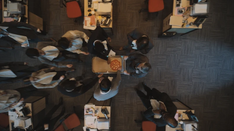 Overhead shot of diverse office workers gathering around a pizza in a box, set on a wheeled cart in a modern office setting.