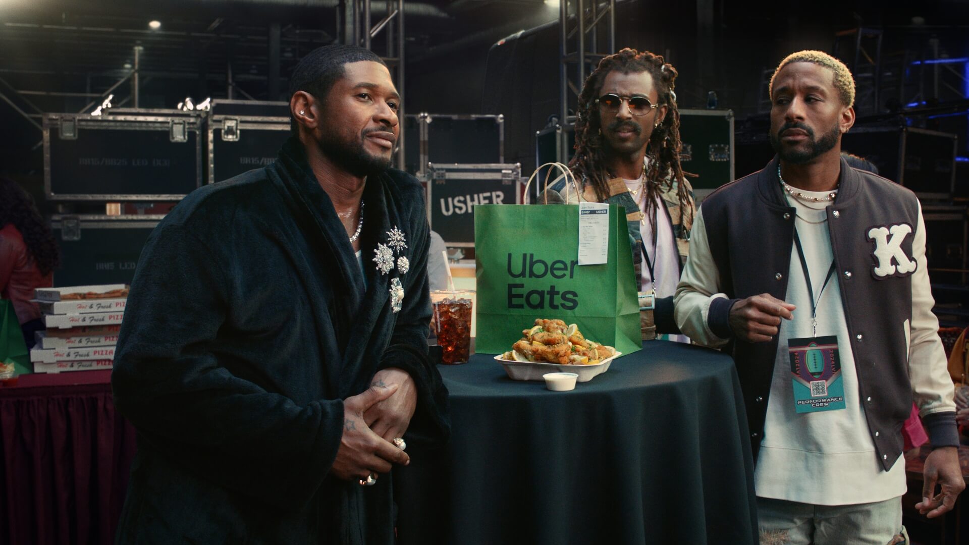 Usher with two men at a table covered in a black tablecloth with food and a drink, in an indoor environment with equipment in the background.