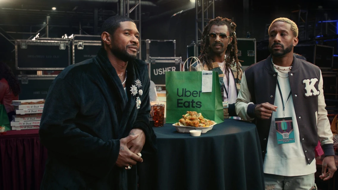 Usher with two men at a table covered in a black tablecloth with food and a drink, in an indoor environment with equipment in the background.