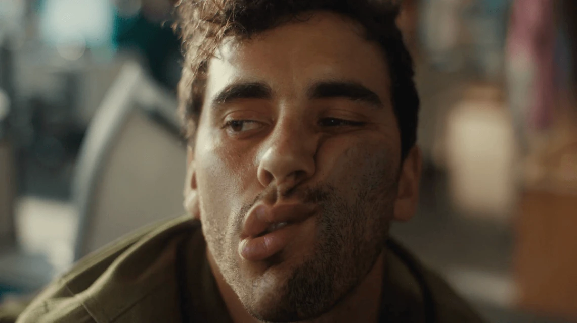 Close-up shot of a young adult male with a slightly dirty face, wearing an olive green jacket, pulling a funny face with his lips puckered.
