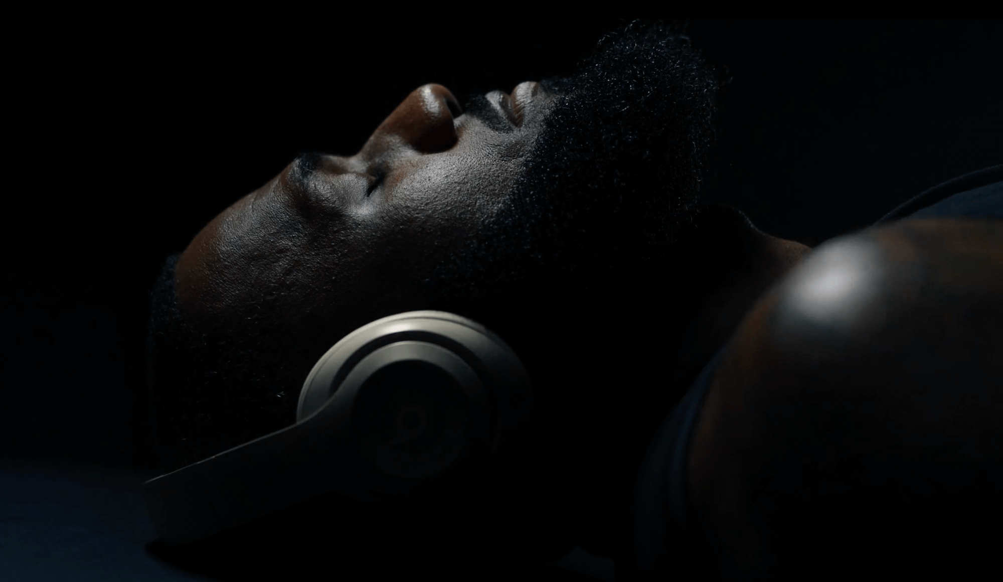 Close-up of African-American man lying down wearing silver headphones against a dark background with dramatic lighting. Low key photo.