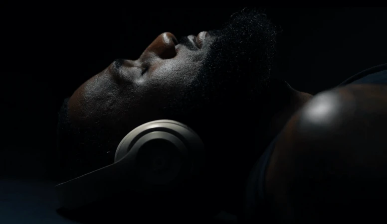 Close-up of African-American man lying down wearing silver headphones against a dark background with dramatic lighting. Low key photo.