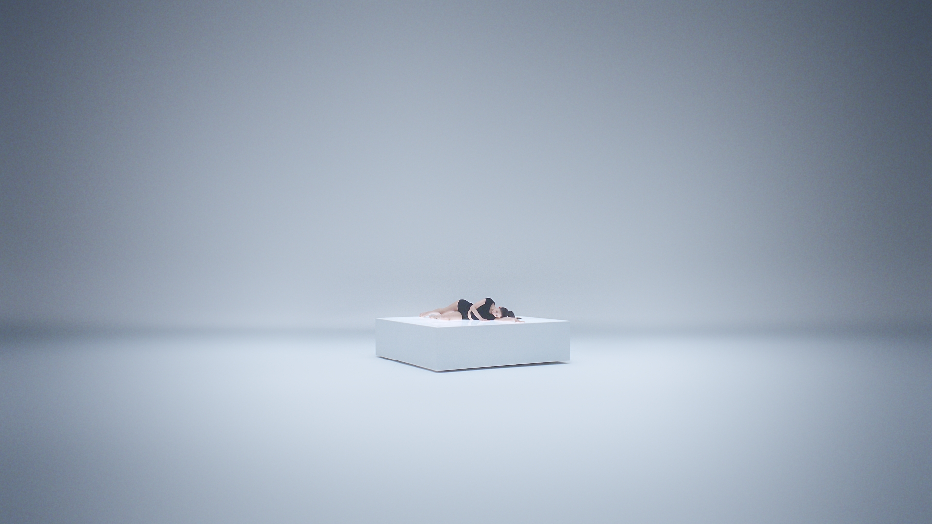 A person lying curled up on a raised white rectangular block in a minimalist grey studio space.