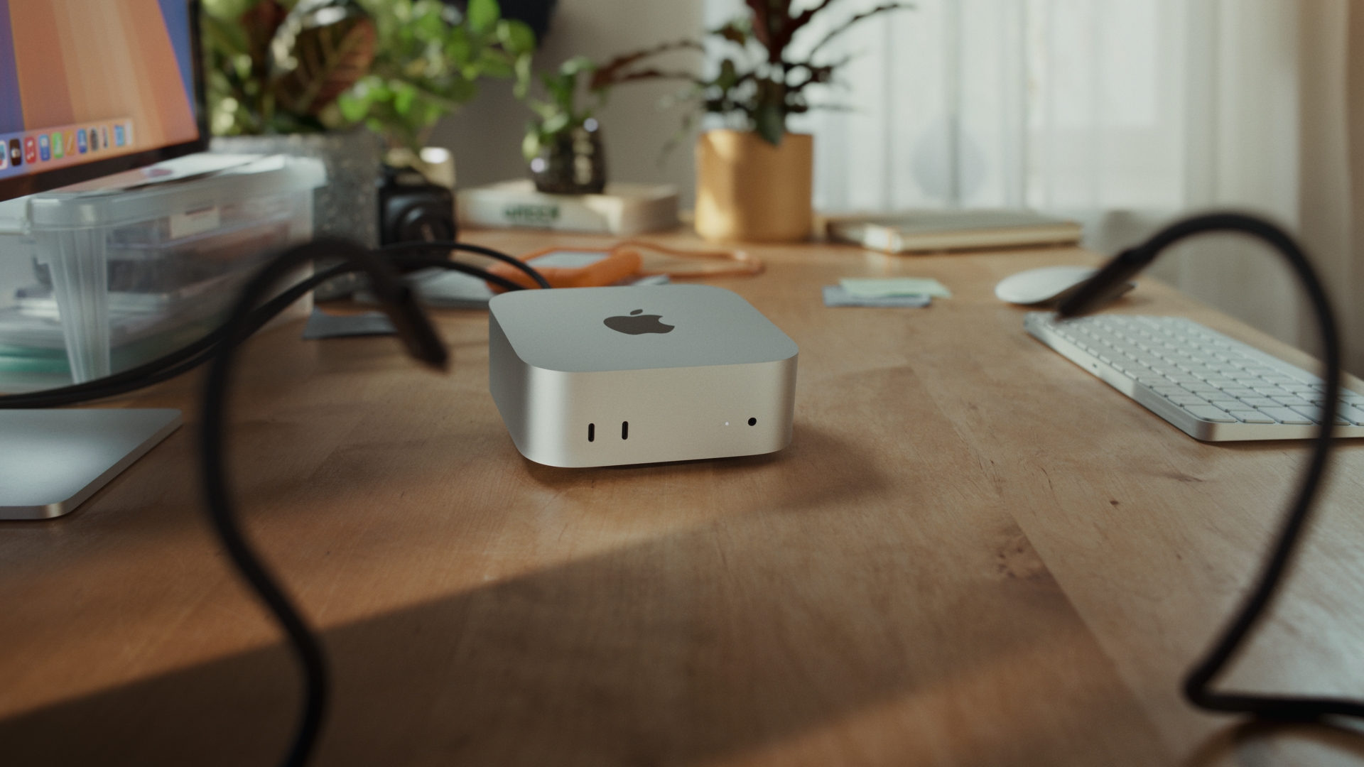Apple Mac Mini M2 on a wooden desk with a keyboard, computer monitor, plants, and other accessories in a brightly lit room.