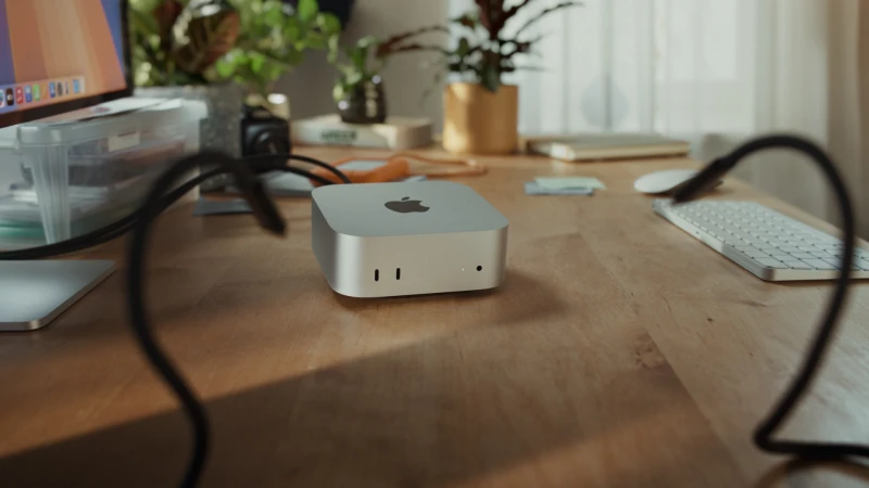 Apple Mac Mini M2 on a wooden desk with a keyboard, computer monitor, plants, and other accessories in a brightly lit room.