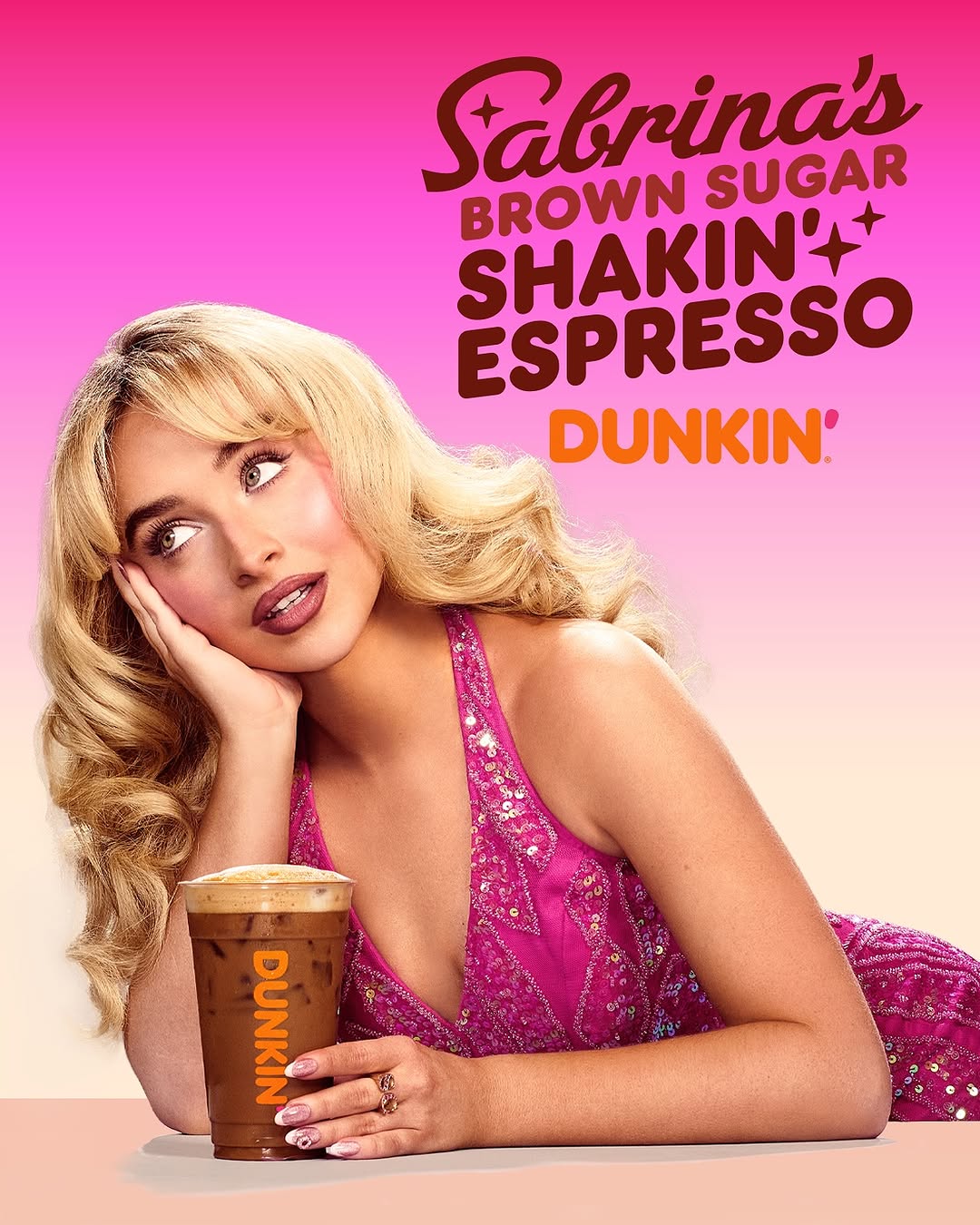 A young woman with blonde hair poses with a Dunkin' iced drink, wearing a pink dress, against a gradient pink background with Dunkin' logo.
