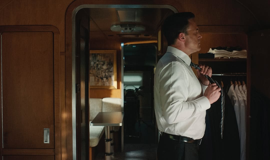 Adult male adjusts necktie while standing in interior space with table, upholstered bench, and a closet of shirts. Interior lighting.