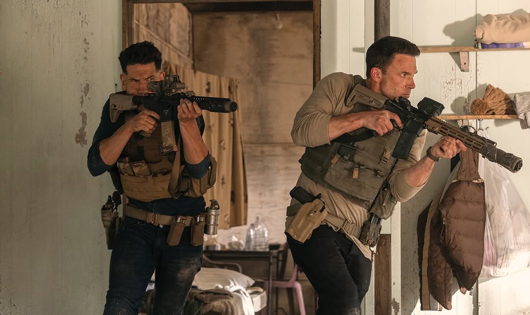 Two adult males in tactical gear hold rifles inside a rustic building, creating a dramatic and intense scene with natural lighting.