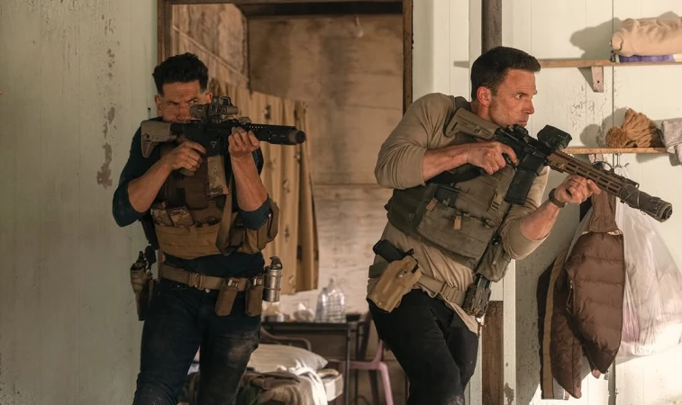 Two adult males in tactical gear hold rifles inside a rustic building, creating a dramatic and intense scene with natural lighting.