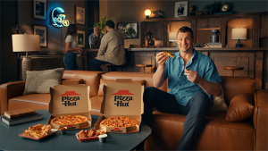 An adult male in a blue shirt sits on a brown leather couch beside Pizza Hut boxes with pizza, in a room with warm lighting.