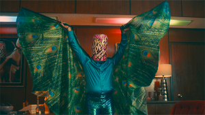 Person in turquoise shirt and sequined pants wearing a Dr. Pepper can on their head and peacock feather cape, indoors with warm lighting.