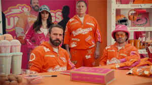 A group of five adults in orange Dunkin' Donuts-themed tracksuits pose at a table with food and beverages inside a Dunkin' store.