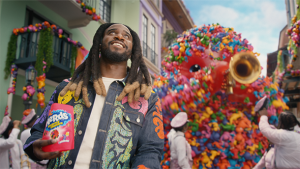 Man in a patterned jacket smiling while holding a box of candy during a street parade with a colorful costumed figure and other people.