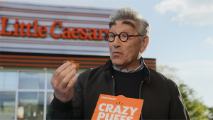 Man holding a Crazy Puff snack from Little Caesars, standing in front of the restaurant on an overcast day in an eye-level shot.