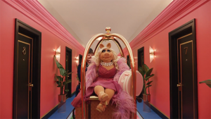Miss Piggy sitting on a luggage cart in a hotel hallway with pink walls and blue carpet. The Muppet is wearing a pink dress.