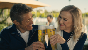 Two adult people, a male and a female, are raising glasses of Michelob Ultra beer in a toast, smiling outdoors during daylight.