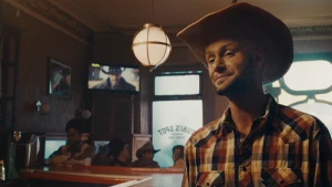 Man in a cowboy hat smiles inside a bar, wearing a plaid shirt with TV and patrons visible in the warm, vintage-lit interior.