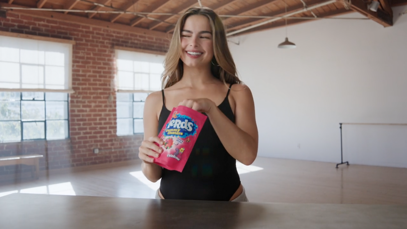 Smiling woman holds NERDS Gummy Clusters candy in an airy loft studio with brick walls and large windows, bathed in soft natural light.