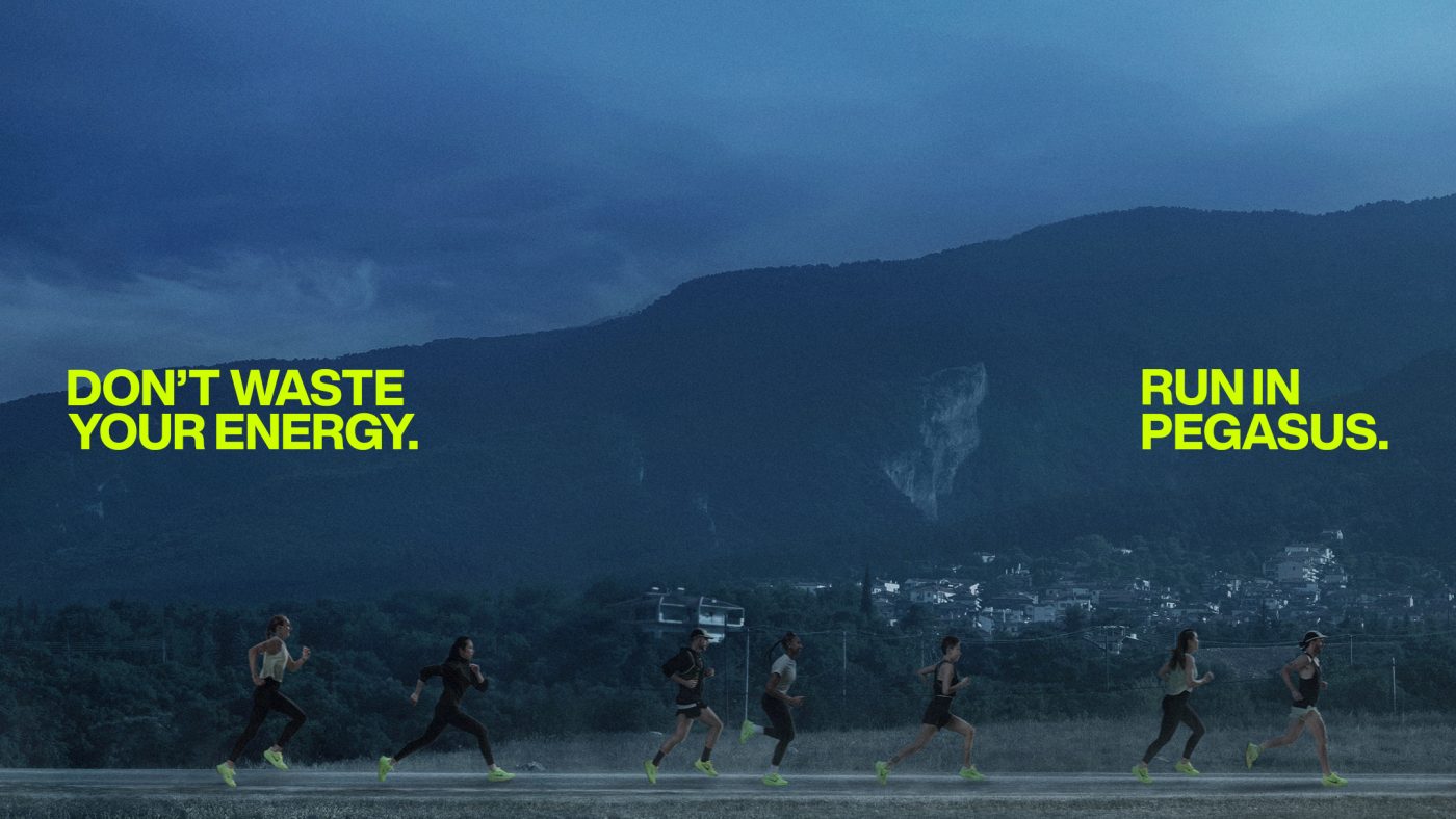 Several people run together outdoors on a track in athletic wear with mountains and the night sky in the background.