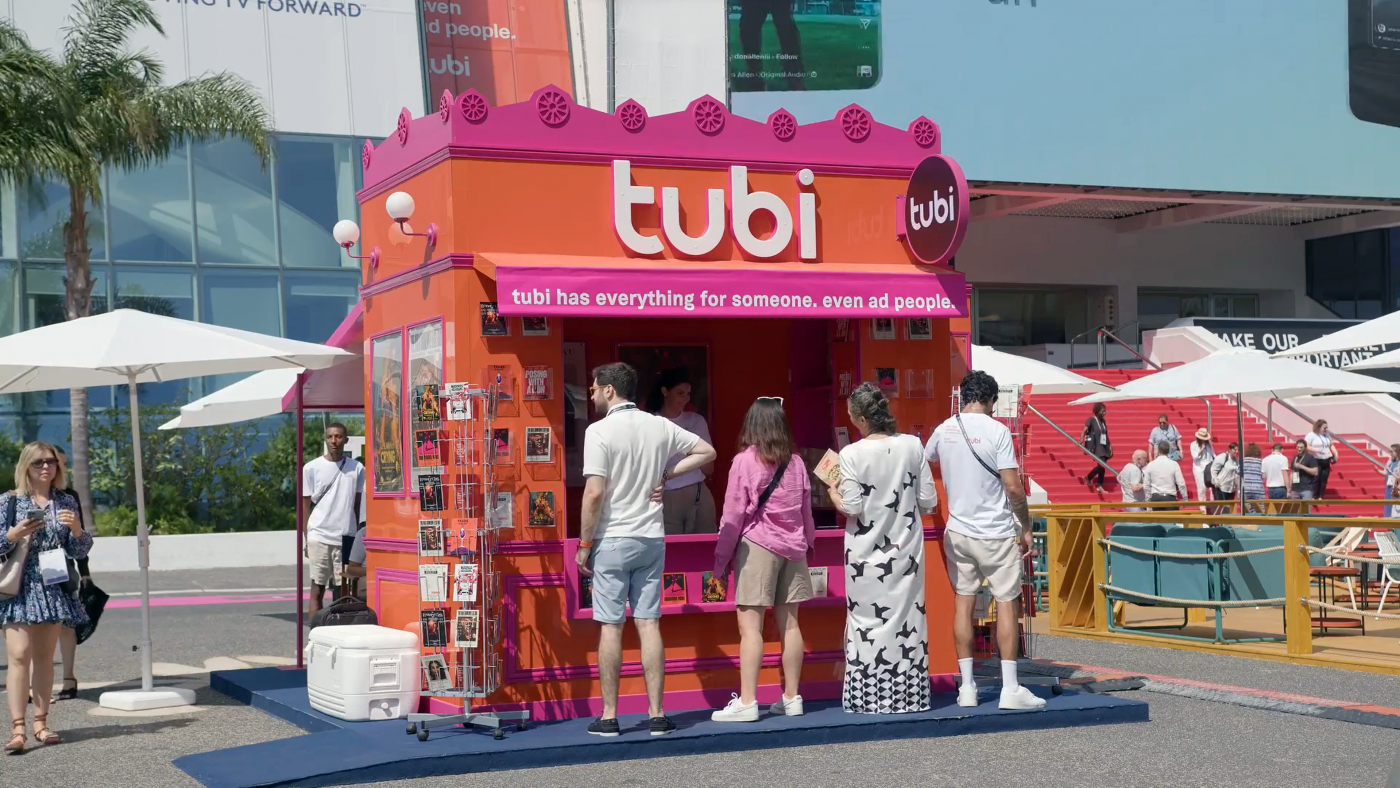 Outdoor shot of a pink and orange Tubi advertising booth, with a few people standing nearby on a sunny day near a red staircase.