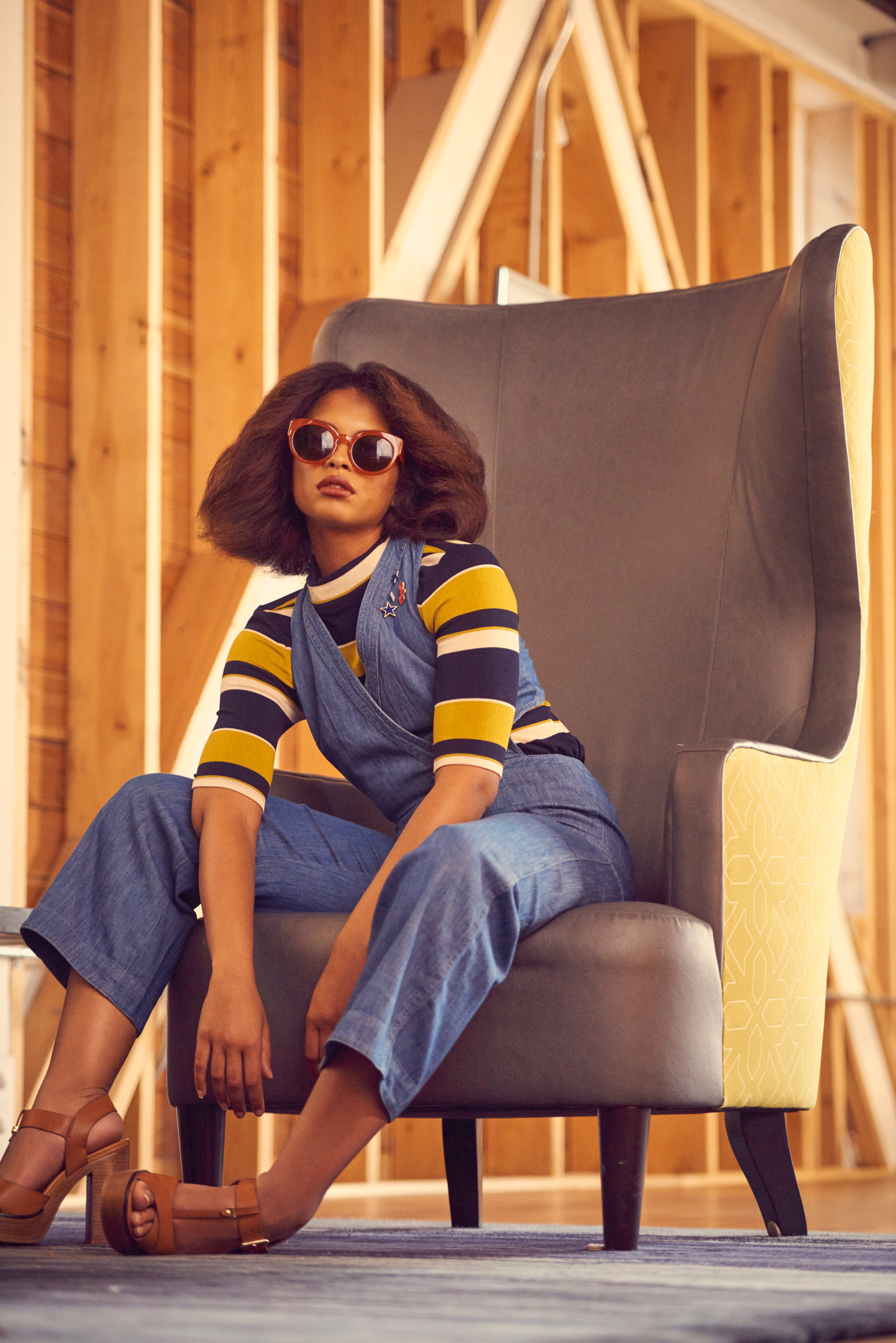 A woman with sunglasses and a denim jumpsuit sits in an armchair against a wood-paneled wall, photographed with bright studio lighting.