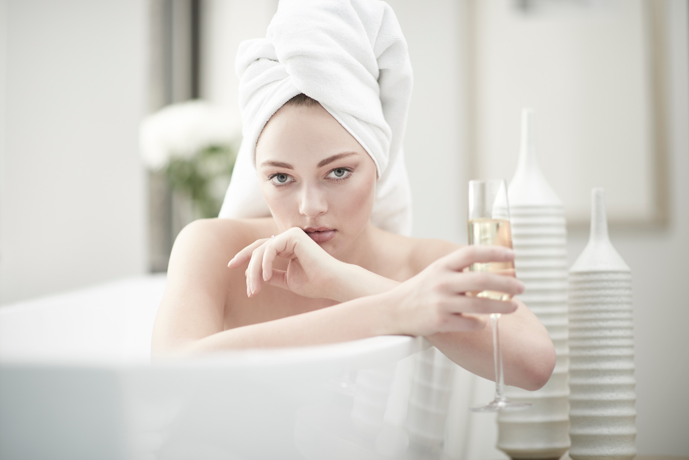 An adult woman with a towel wrapped around her head is sitting in a bathtub while holding a glass of champagne in a bright bathroom.