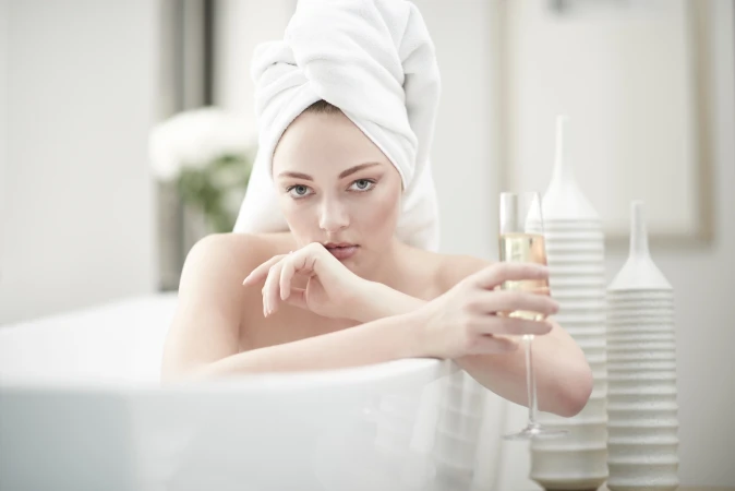 An adult woman with a towel wrapped around her head is sitting in a bathtub while holding a glass of champagne in a bright bathroom.