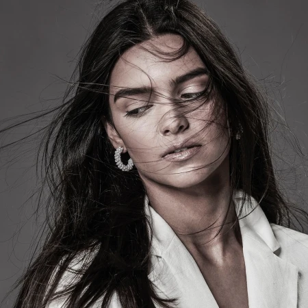Close-up monochrome portrait of a woman with dark hair blowing across her face, wearing pearl hoop earrings and a white jacket.