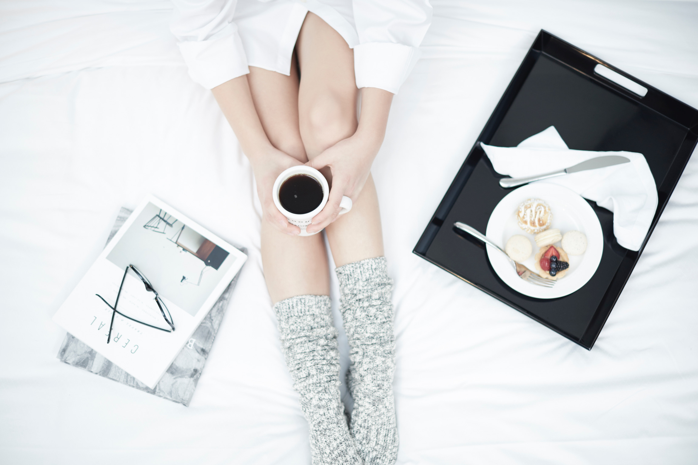Woman sitting on bed holding coffee with breakfast tray and magazine with glasses, suggesting a relaxed morning routine.
