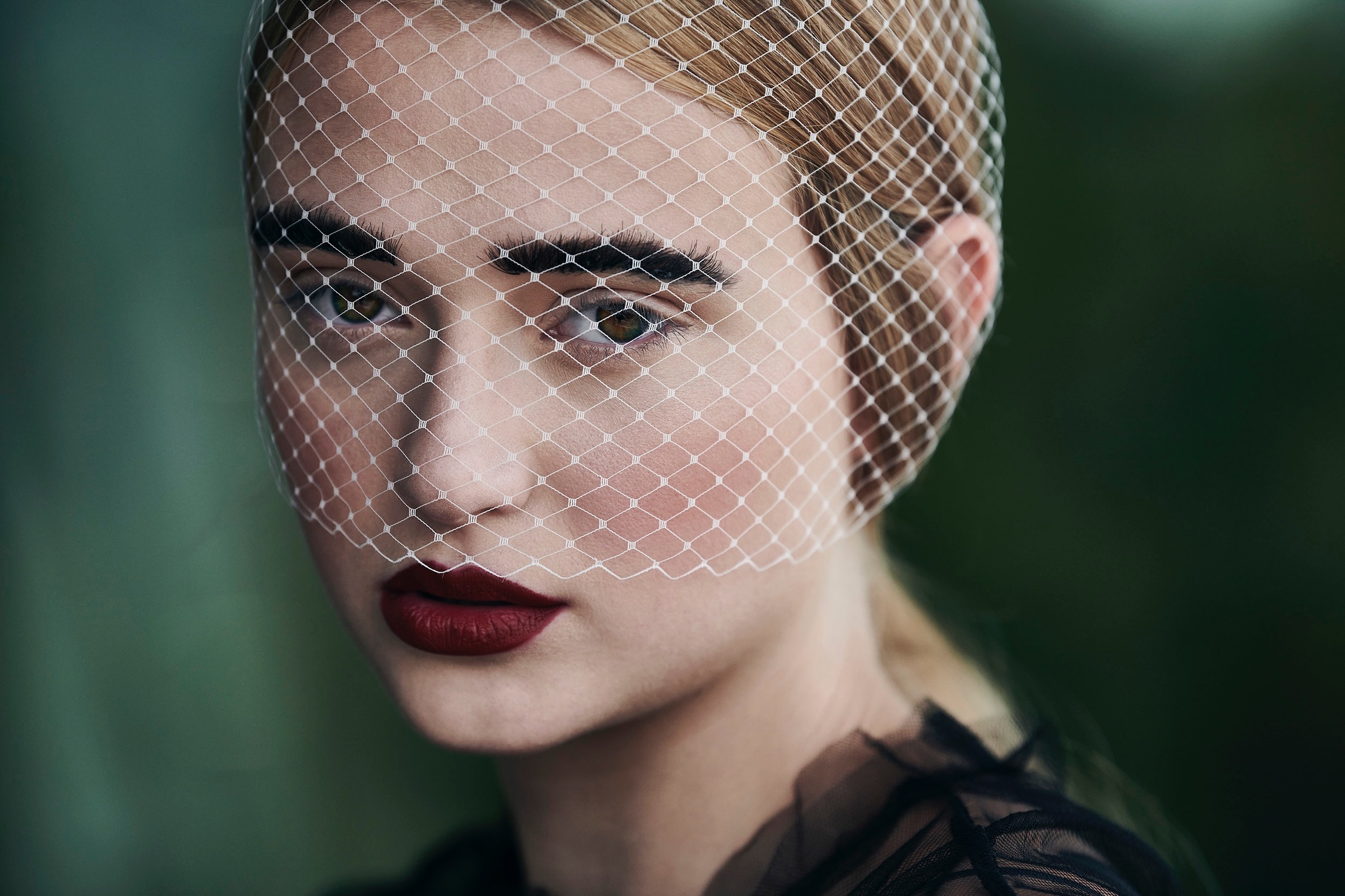 Close-up portrait of a fair-skinned young woman with dark red lipstick and a netting veil against a blurred green background.