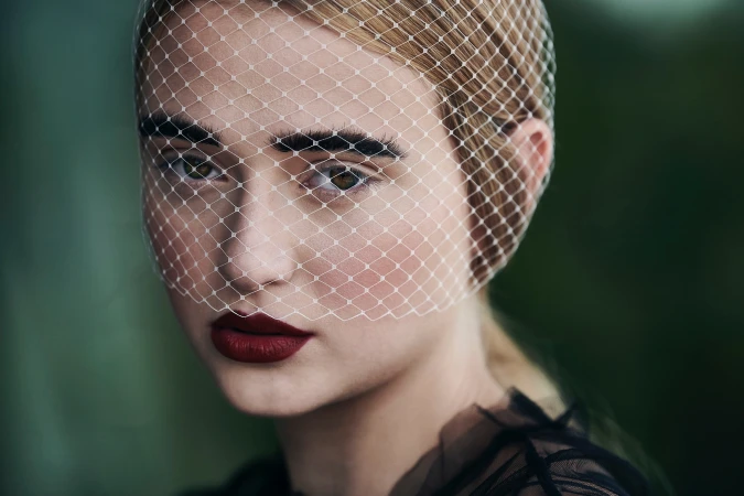 Close-up portrait of a woman wearing a white mesh veil over her face and dark lipstick by photographer Ken Friberg.