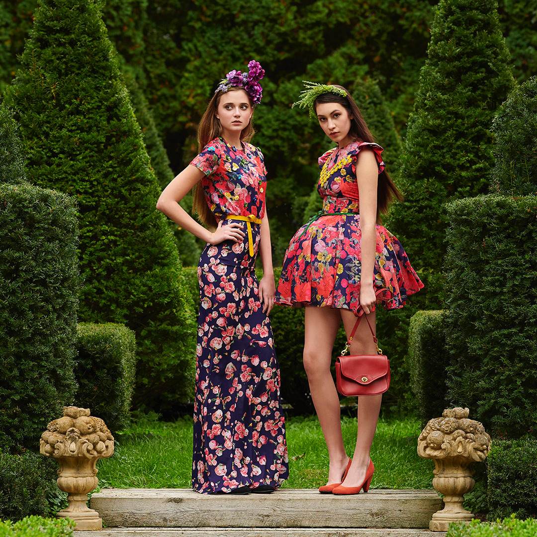 Two women in floral print dresses stand on stone steps in a formal garden with sculpted trees and manicured greenery on a sunny day.