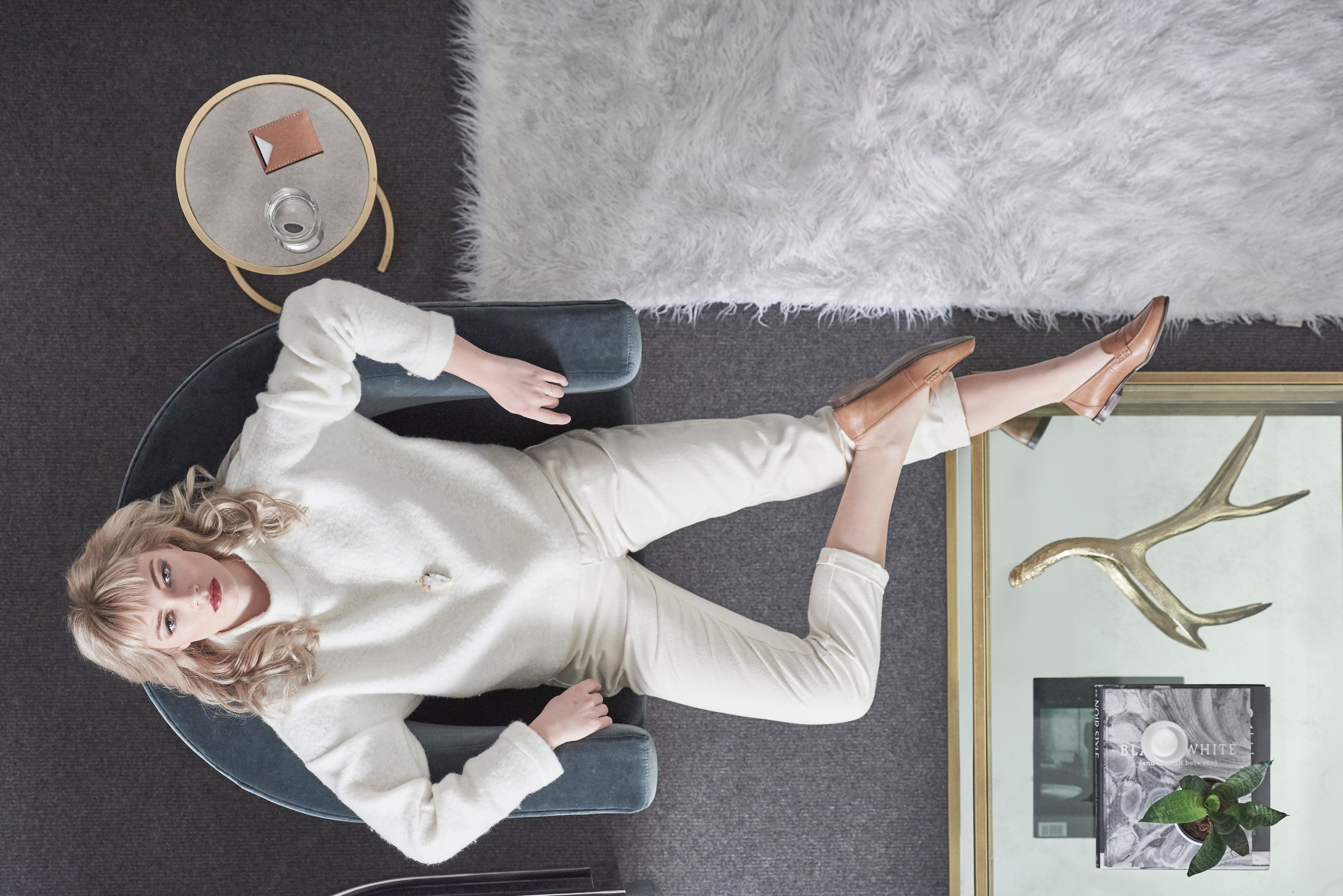 Overhead view of a blonde woman lying on a blue velvet chair in a white sweater, cream pants, and light-brown shoes near a side table.