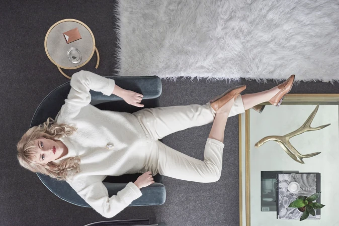 Overhead view of a blonde woman lying on a blue velvet chair in a white sweater, cream pants, and light-brown shoes near a side table.