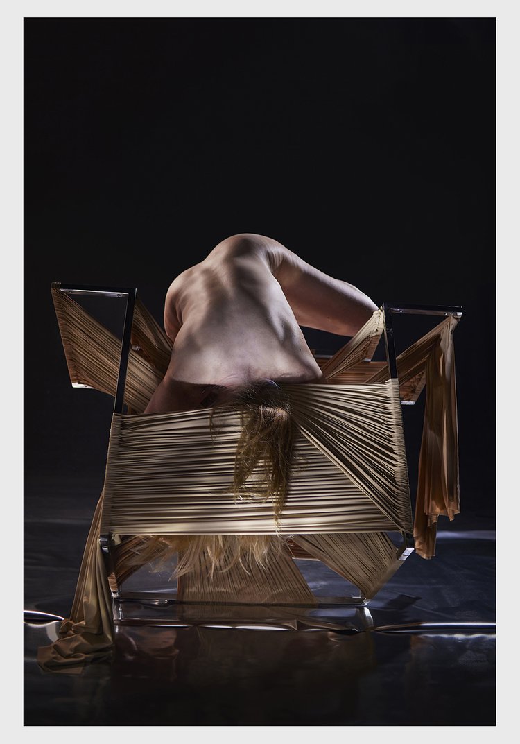 Abstract art photography of an androgynous human figure sitting backwards on a chair wrapped in beige ribbons in a dark studio.