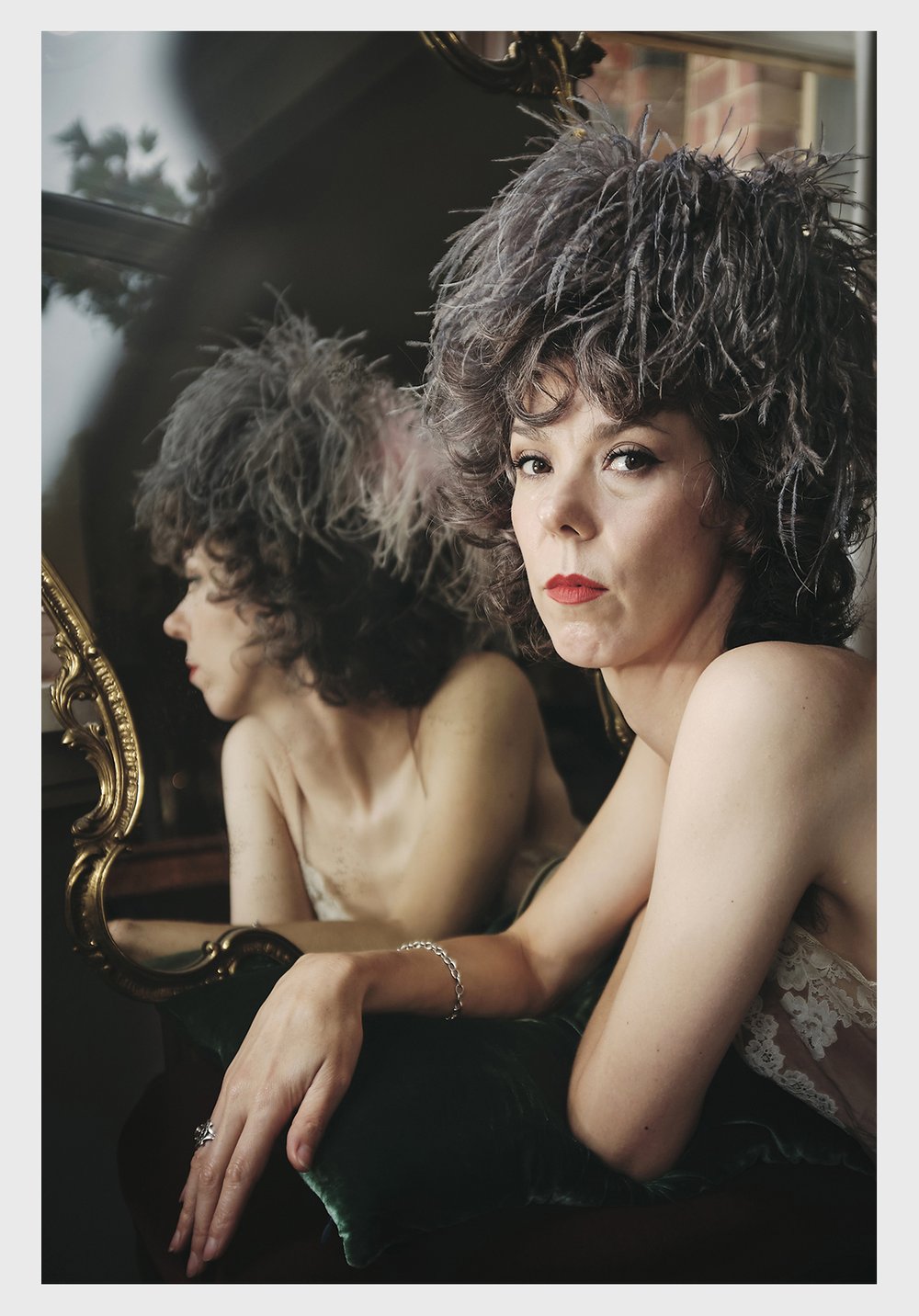 Portrait of a woman with an extravagant feathered hat leaning on a green velvet surface with a mirror reflecting her image indoors.