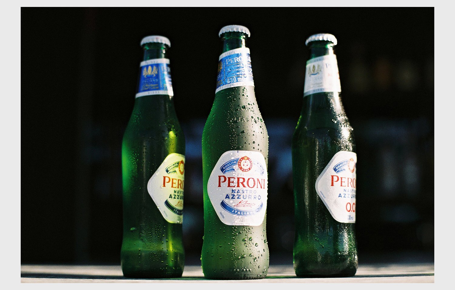 Three green glass Peroni Nastro Azzurro beer bottles sit on a surface, featuring condensation and white labels against a dark background.