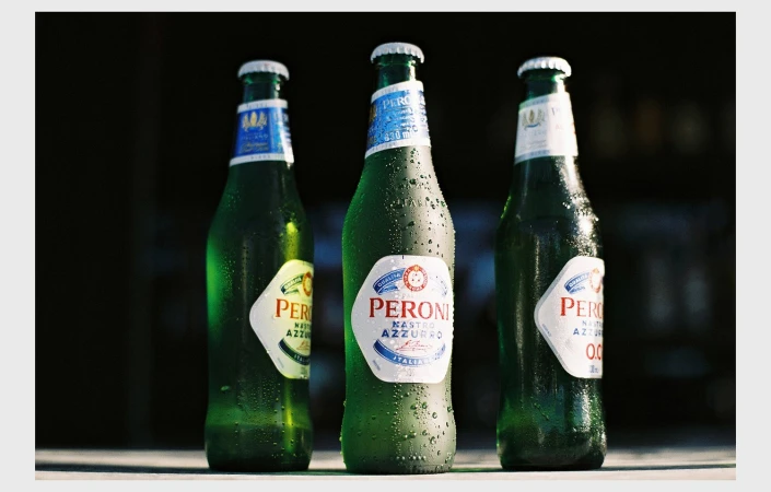 Three green glass Peroni Nastro Azzurro beer bottles sit on a surface, featuring condensation and white labels against a dark background.