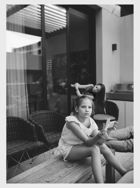 Black and white photo of a young girl sitting on a bench with an older girl in the background near window blinds.