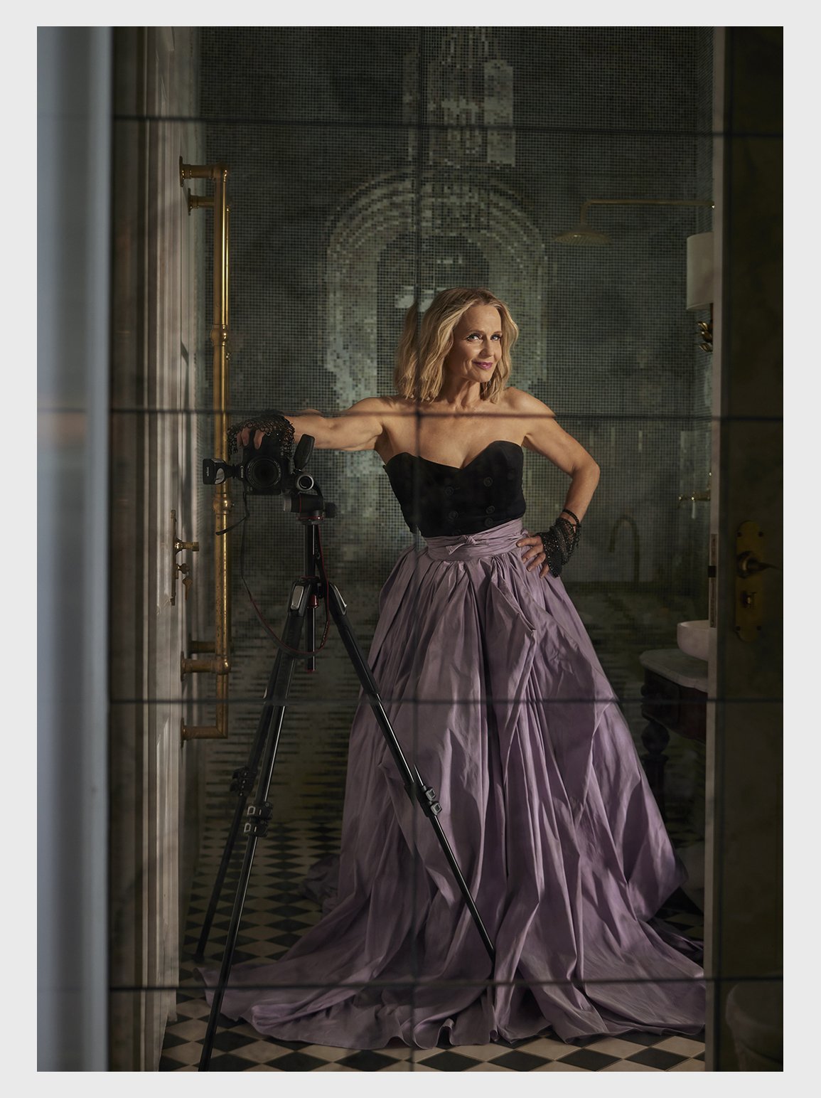 Woman in black top and lilac ballgown poses indoors with professional camera on a tripod in front of decorative wallpaper.