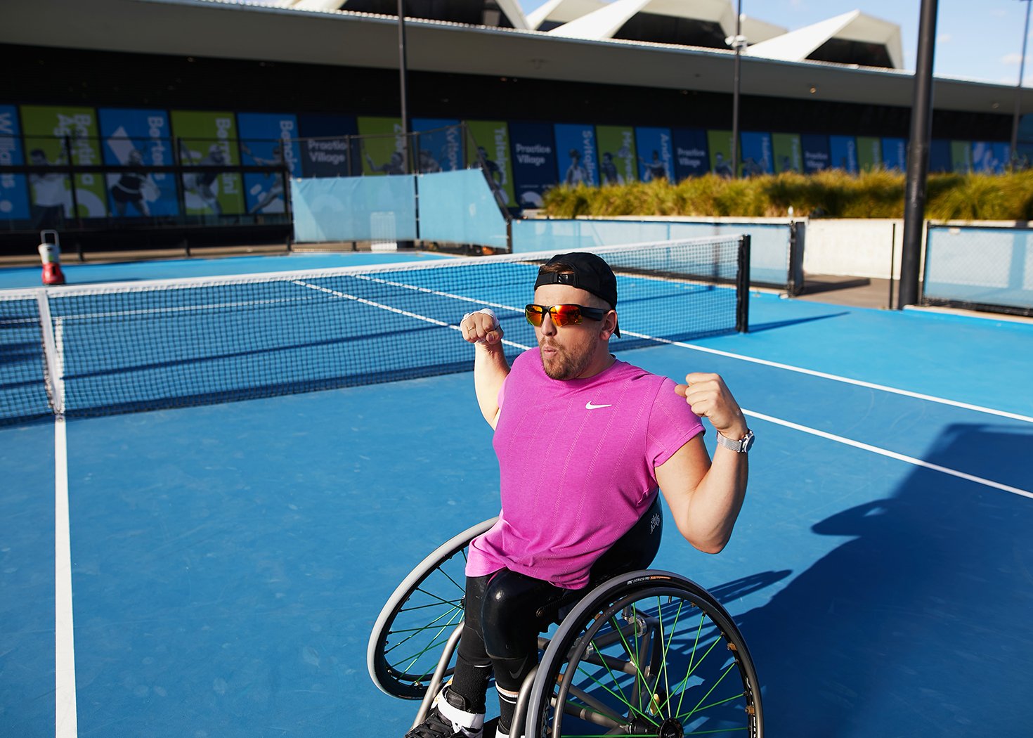 Man in wheelchair posing on tennis court with arms raised. He's wearing sunglasses, cap, pink t-shirt with net and building background.